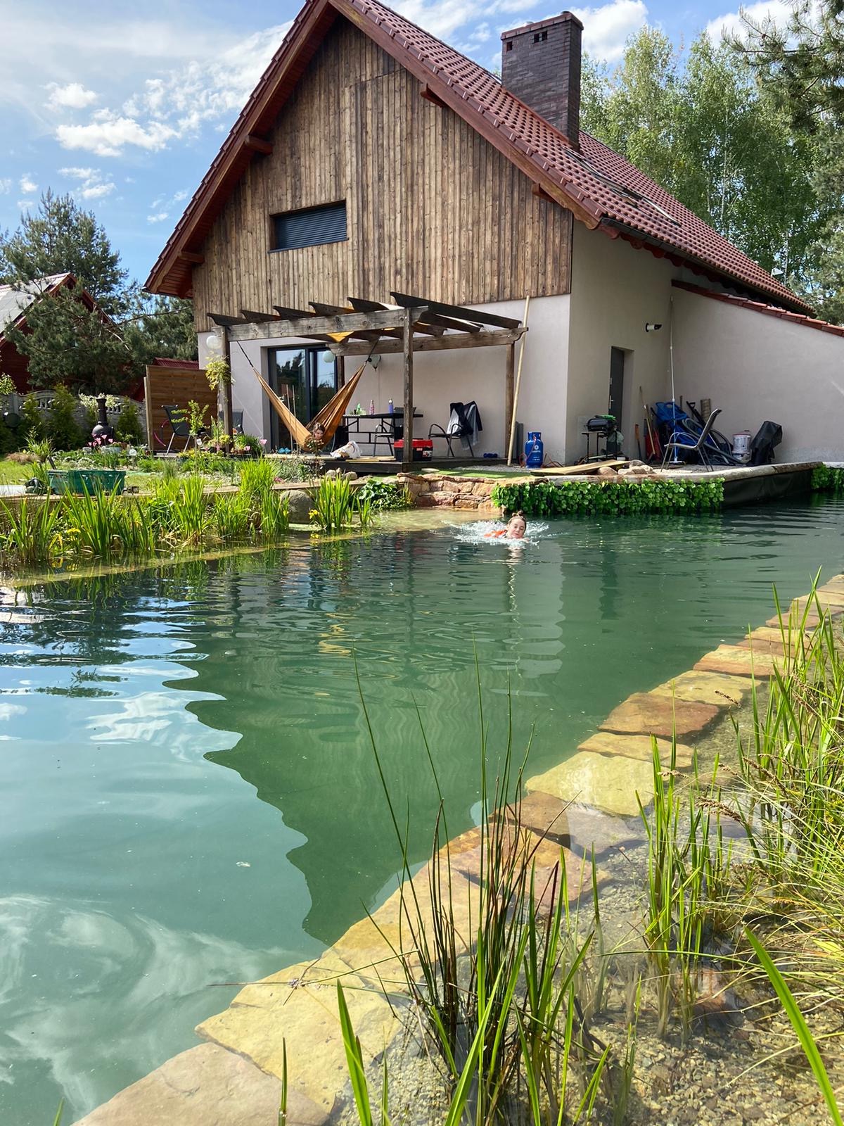 Naturalny basen w ogrodzie z drewnianym domem w tle, osoba pływająca w wodzie, roślinność wodna, pergola z hamakiem.