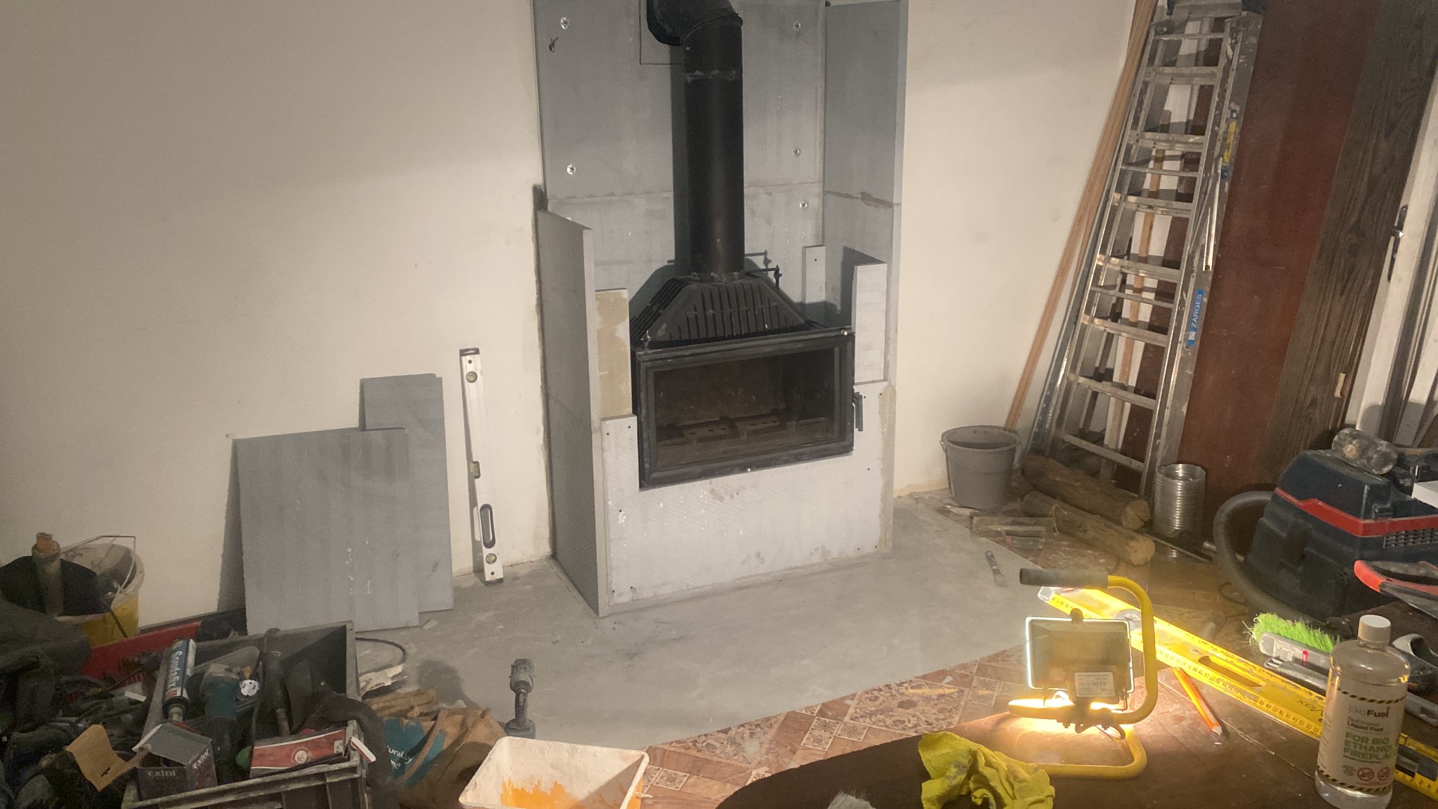 Montaż wkładu kominkowego z czarną obudową i rurą dymową, obudowany płytami gipsowymi, widoczne narzędzia i drabina w tle.