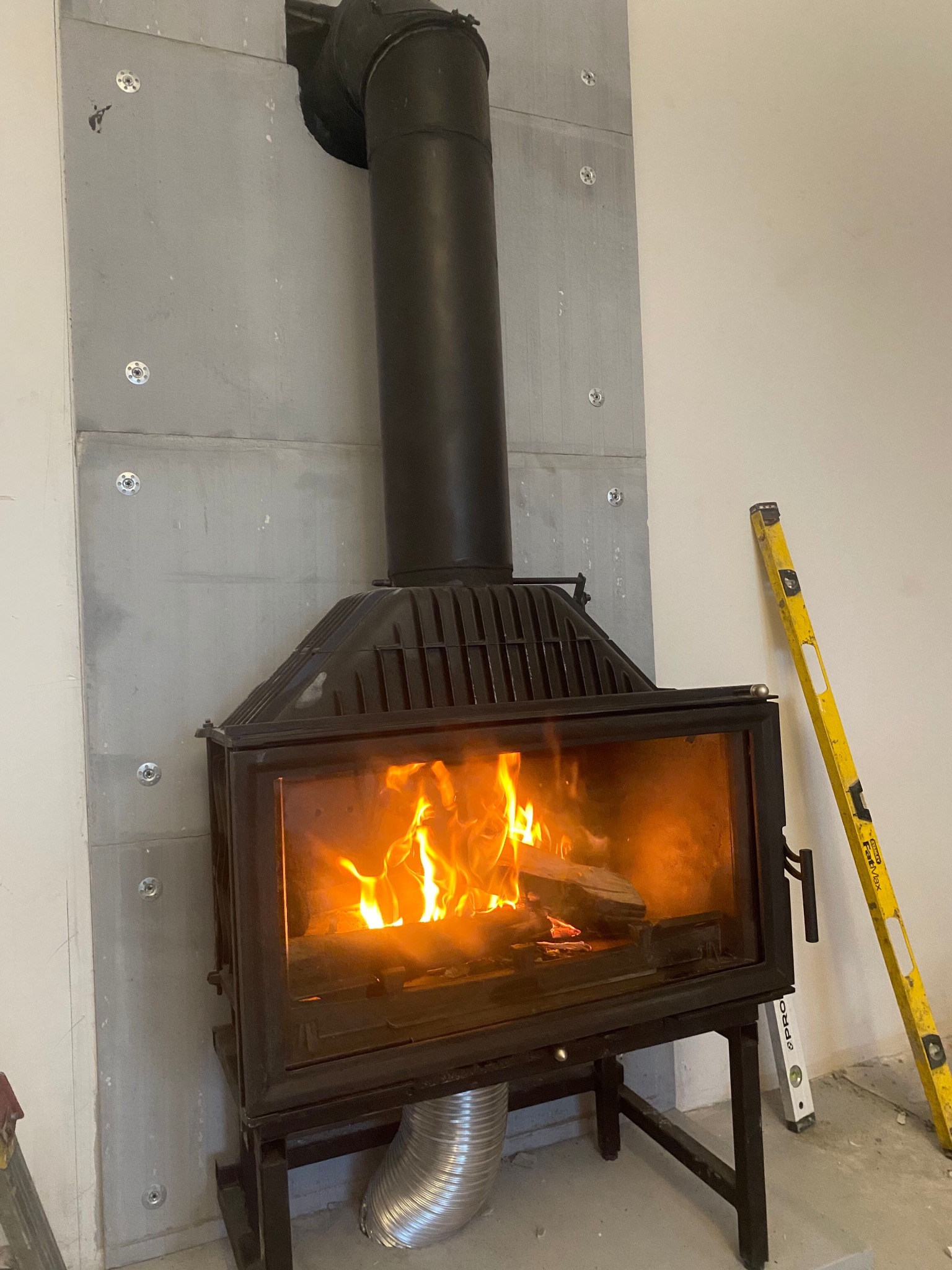 Wnętrze z zamontowanym czarnym kominkiem z płomieniami, obudowanym szarymi płytami przymocowanymi metalowymi śrubami, widoczna poziomica oparta o ścianę.