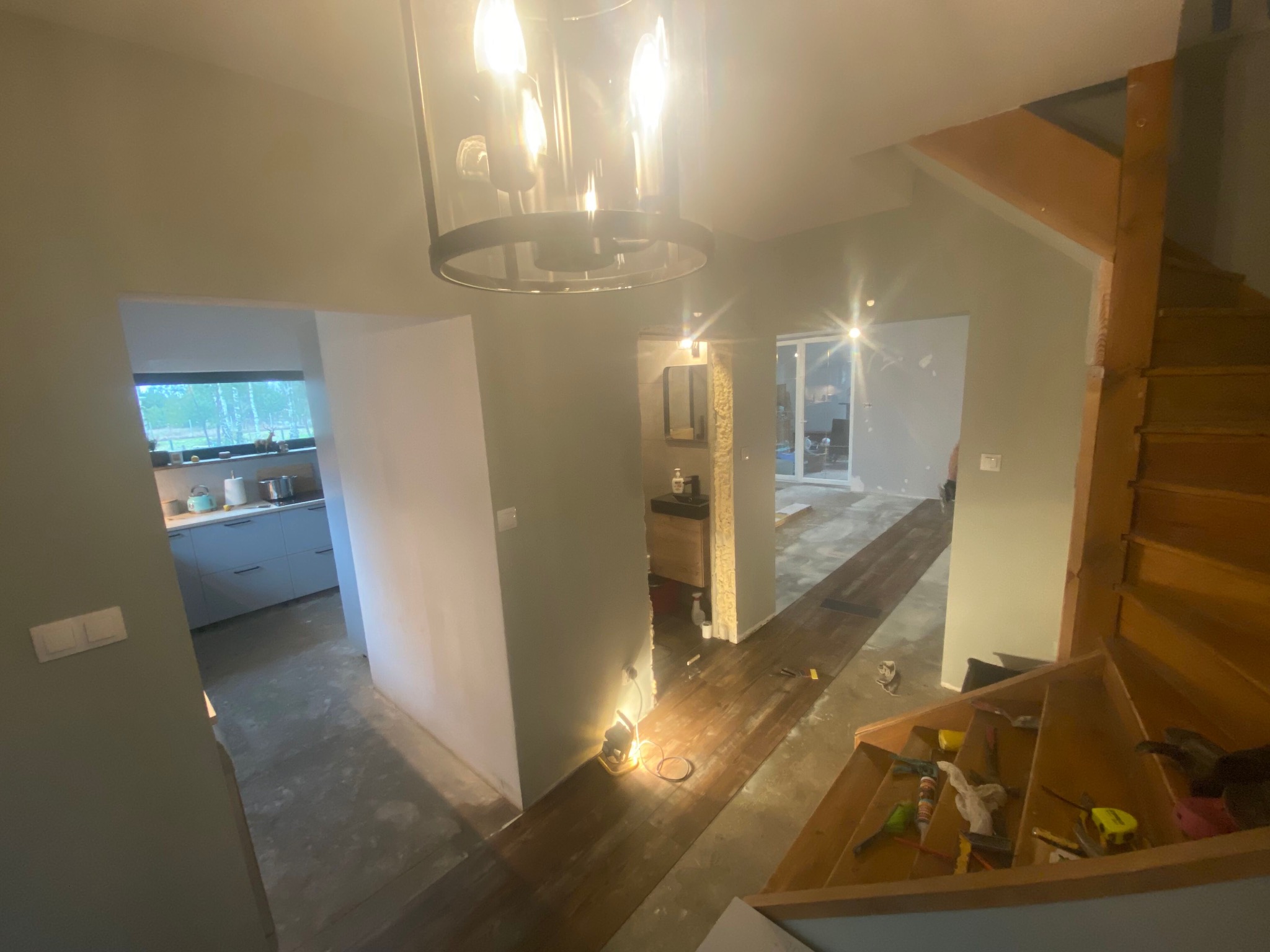 Wnętrze domu w trakcie remontu, widoczne schody drewniane, fragment kuchni z niebieskimi szafkami, korytarz z przygotowaną podłogą i lampą budowlaną oraz łazienka w tle. Ściany w szarym odcieniu...