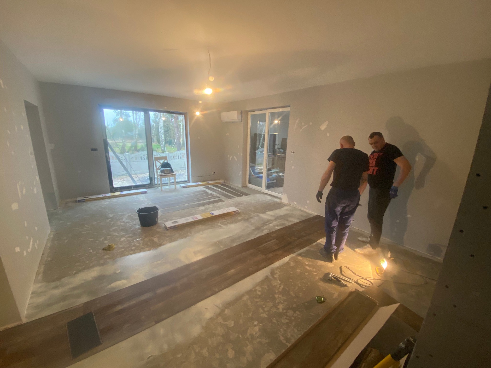 Dwóch pracowników w trakcie układania paneli podłogowych w jasnym, surowym wnętrzu z widocznymi śladami szpachlowania na ścianach i przygotowaną podłogą. Na podłodze leżą panele, narzędzia i wiadro.