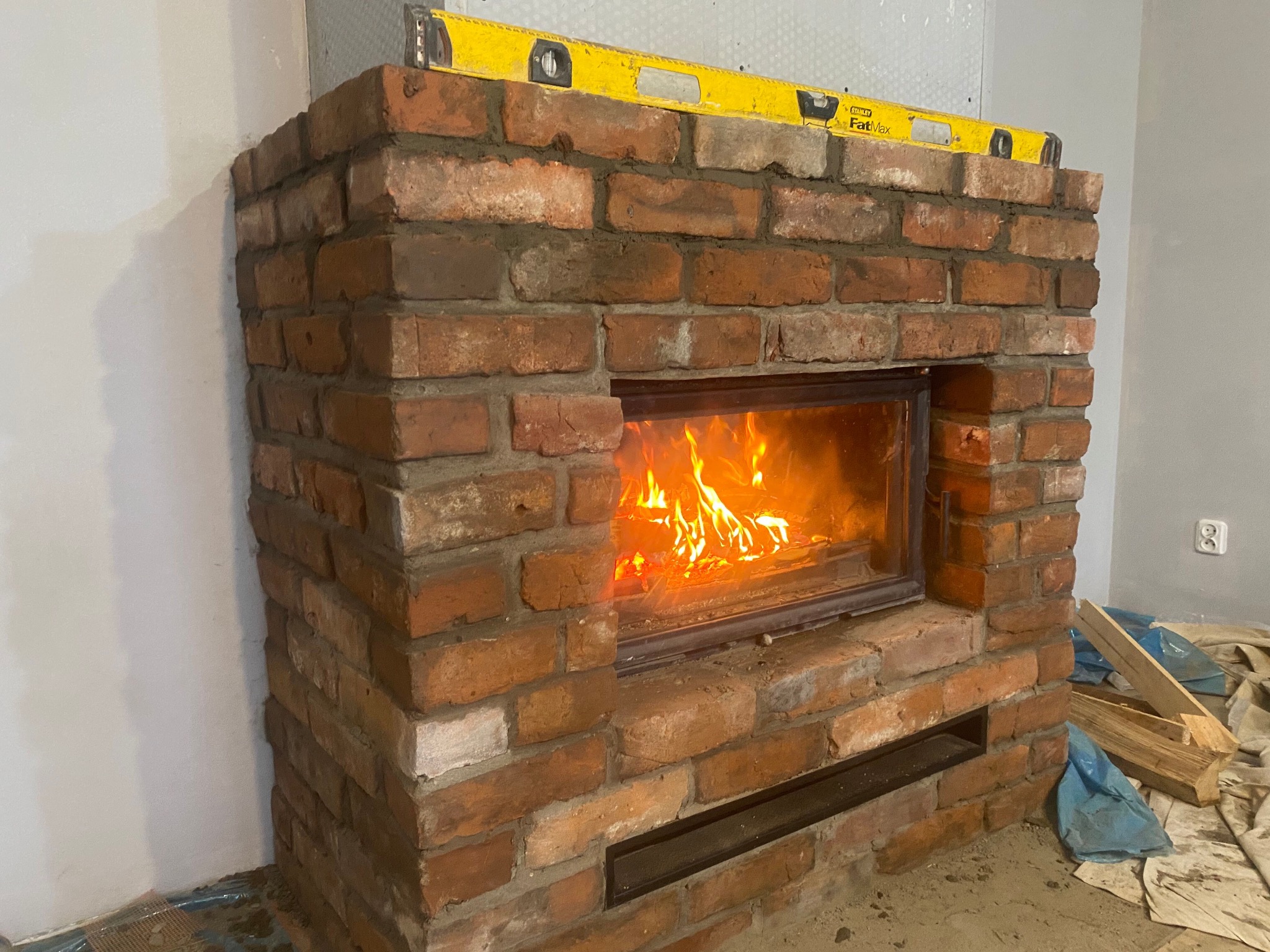 Murowany kominek z czerwonej cegły z płomieniami wewnątrz paleniska, poziomica na górze, w trakcie budowy, ściany w tle przygotowane pod malowanie.