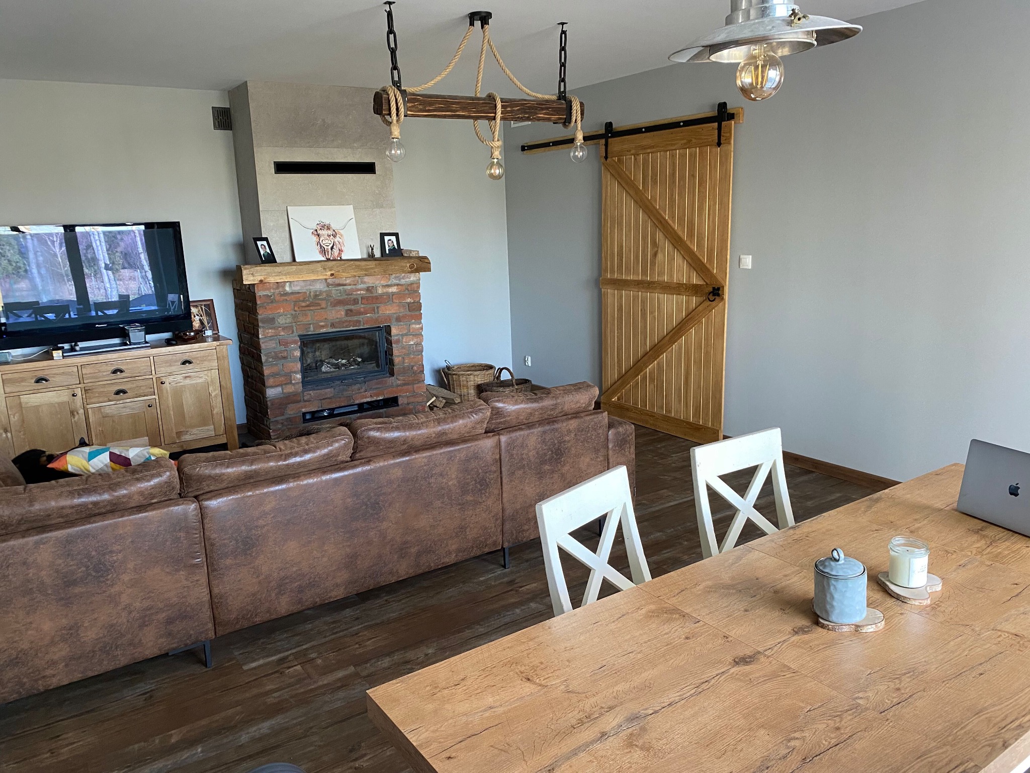 Jasne wnętrze salonu z ceglanym kominkiem, drewnianą podłogą i przesuwnymi drzwiami stodołowymi, oświetlone lampą w stylu rustykalnym z linami, z widocznym fragmentem stołu z białymi krzesłami...