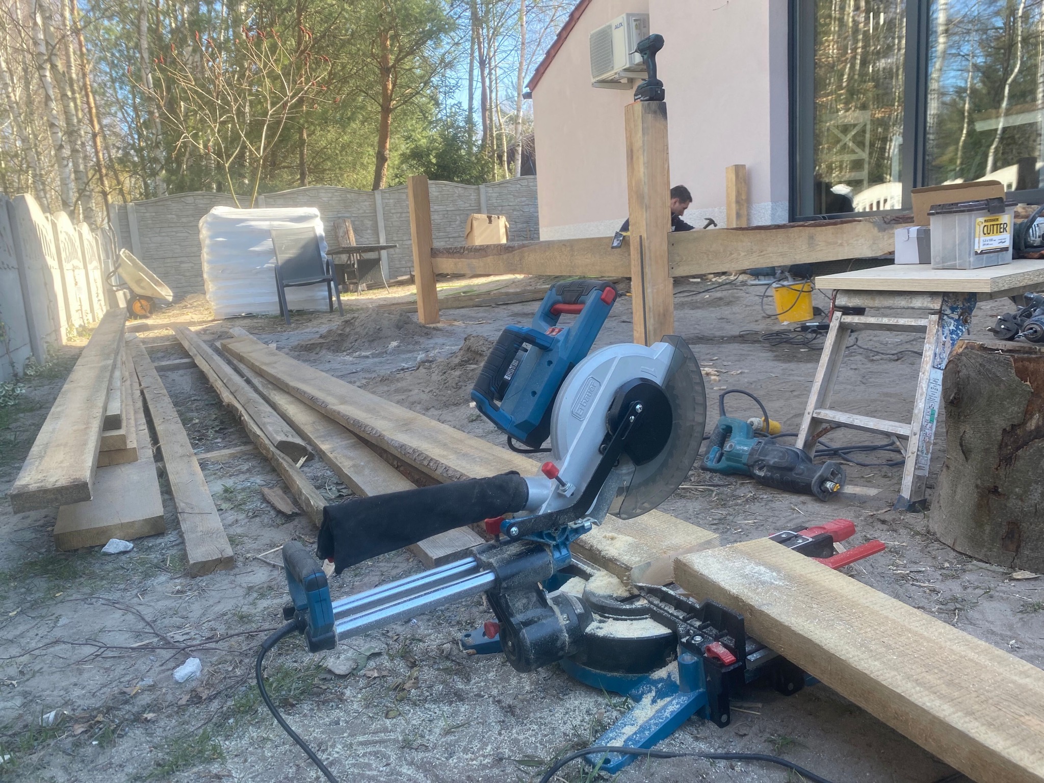 Budowa drewnianego tarasu: piła ukośnica tnie deski, w tle konstrukcja z belek i pracownik z młotkiem.