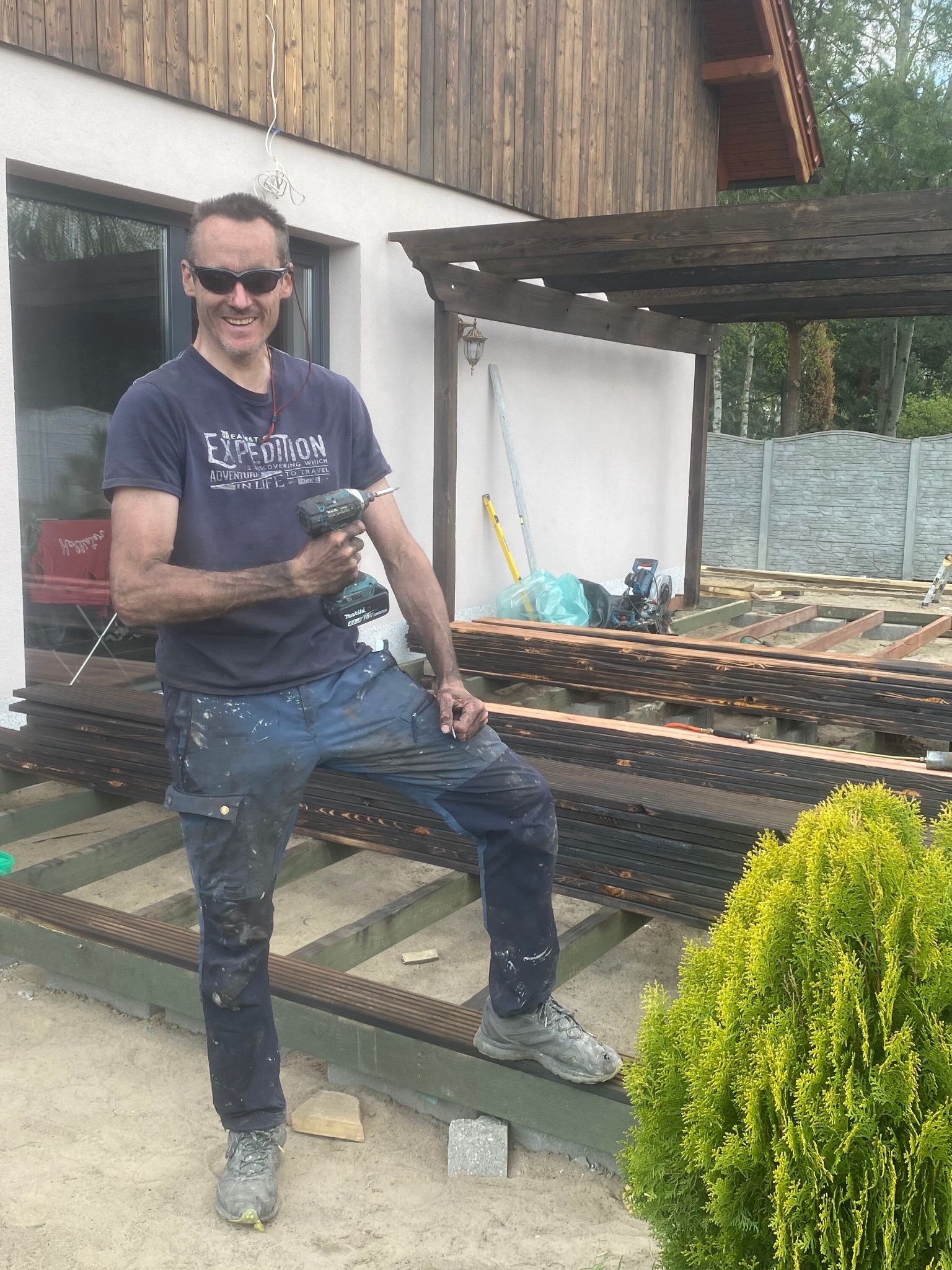 Uśmiechnięty mężczyzna w okularach i roboczym stroju, trzymający wiertarko-wkrętarkę Makita, pozuje na tle budowanej drewnianej konstrukcji tarasu z pergolą, z widocznymi deskami, piłą i poziomnicą.