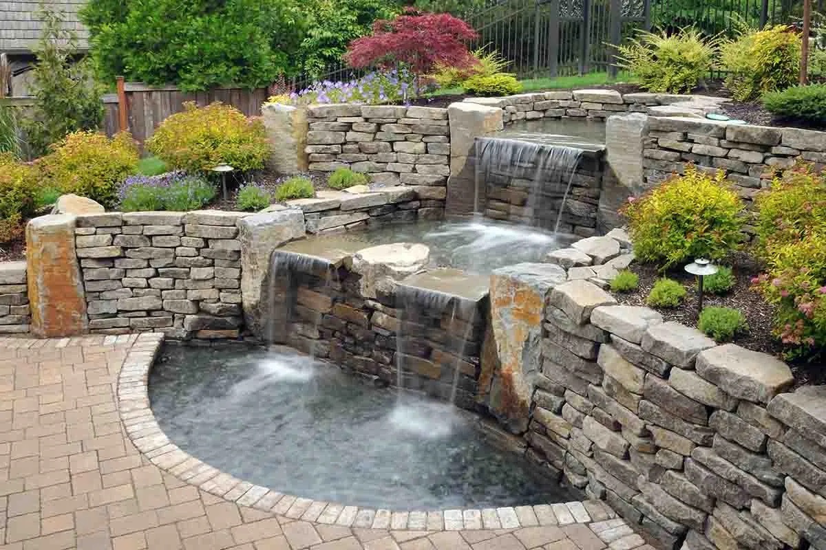 Trójpoziomowy, kamienny wodospad ogrodowy z kaskadami, otoczony roślinnością i brukowanym patio, z widoczną czerwoną japońską mapą w tle.