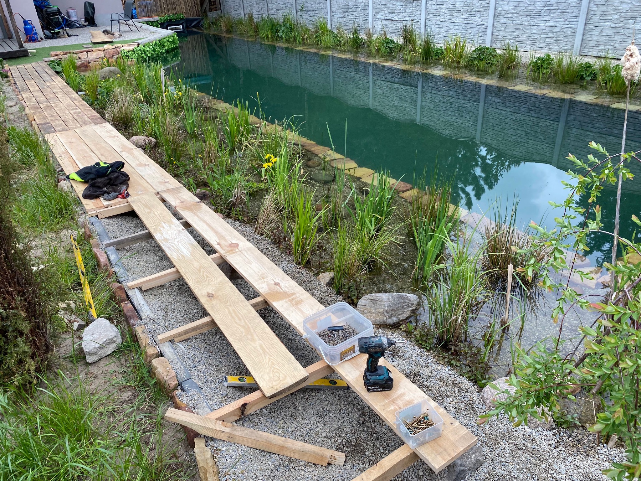 Budowa drewnianego pomostu nad stawem: deski, poziomica, wkrętarka Makita i pojemniki z wkrętami na tle wodnej roślinności.
