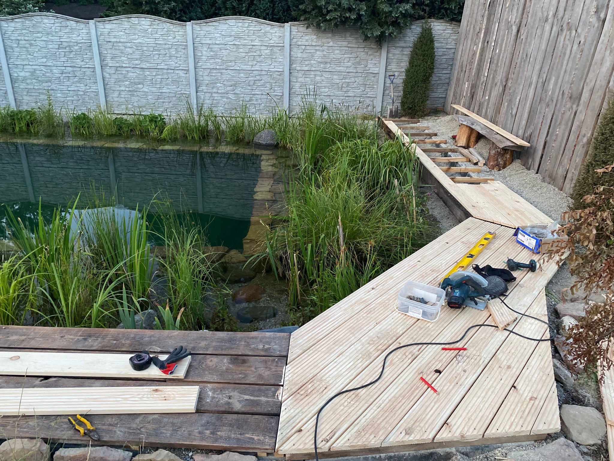 Budowa drewnianego tarasu nad oczkiem wodnym, widoczne narzędzia i materiały budowlane, w tle betonowe ogrodzenie i roślinność.