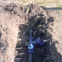 Niebieska armatura na czarnym rurociągu w wykopie ziemnym, widoczne korzenie, w tle fragment opony koparki.