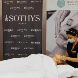 Kosmetolog w maseczce wykonuje masaż twarzy klientce leżącej na białym prześcieradle; w tle banery reklamowe salonu z logo Sothys i ofertą zabiegów.