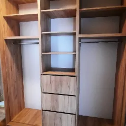 Wnętrze szafy wnękowej z trzema szufladami i półkami, wykończonej w okleinie drewnopodobnej, z widocznym fragmentem dywanu na podłodze.