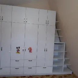 Biała szafa na wymiar z półkami schodkowymi w Lublińcu, ozdobiona naklejkami z postaciami z bajek, widoczna obok brązowych drzwi.