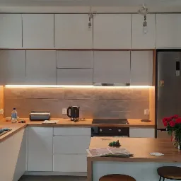 Nowoczesna kuchnia z białymi frontami szafek, drewnianym blatem i oświetleniem LED podszafkowym, uzupełniona o drewniany stół z bukietem czerwonych róż, widok na dekoracyjne oświetlenie ścienne...