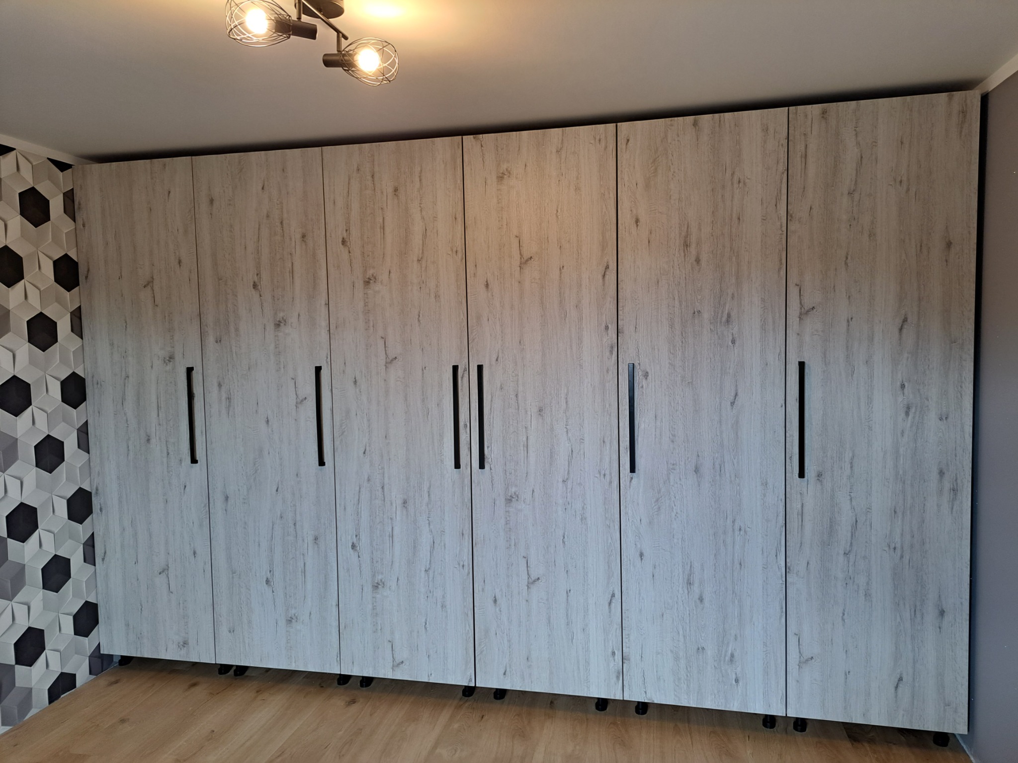 Długa szafa z dziesięcioma drzwiami o wzorze jasnego drewna, wyposażona w czarne, pionowe uchwyty, stoi na małych czarnych kółkach, z widoczną ścianą z geometrycznym wzorem w tle.