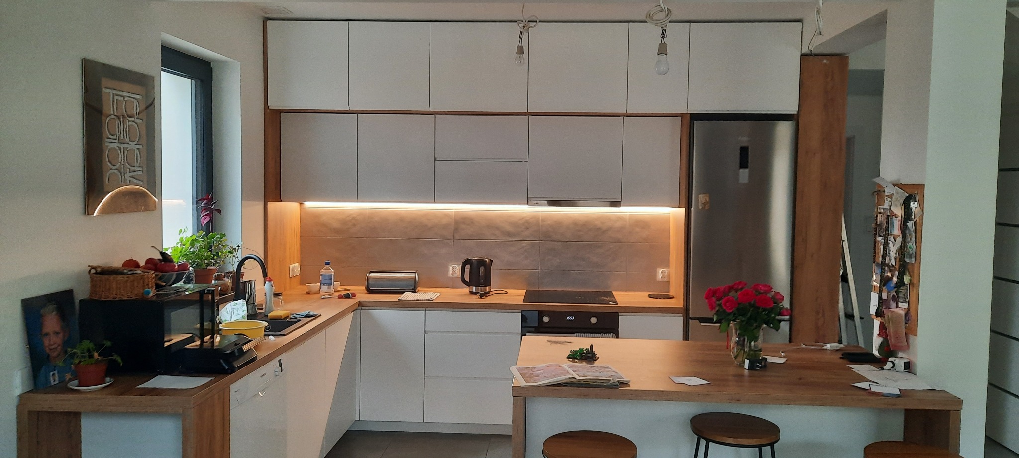 Nowoczesna kuchnia z białymi szafkami, drewnianym blatem i oświetleniem LED pod szafkami, z wyspą kuchenną i dwoma stołkami barowymi, oraz dekoracjami na blacie i ścianie.