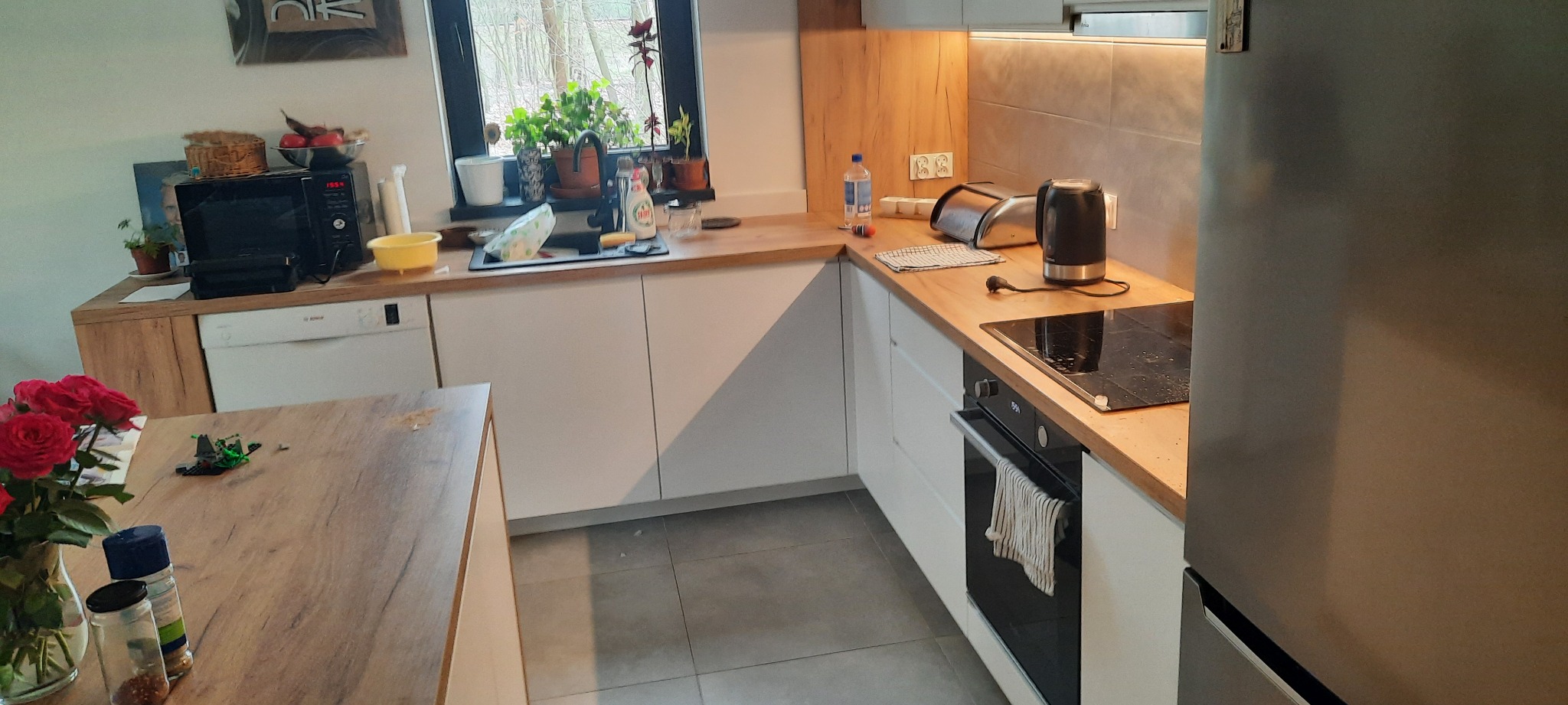 Jasna kuchnia z drewnianymi blatami, widok na okno z roślinami, czajnik elektryczny i płyta indukcyjna na blacie, kwiaty i przyprawy na stole w pierwszym planie.