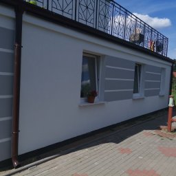 Mateusz Giruć elewacje i ocieplenia - Ściana budynku po tynkowaniu z szarymi poziomymi pasami, brązowa rynna, widoczny fragment balkonu z czarną balustradą i czerwona pompa wodna na kostce brukowej.