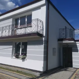 Dwupiętrowy dom z jasną elewacją w odcieniach szarości i bieli, z balkonami o geometrycznych balustradach i kwiatami w oknach, sfotografowany w słoneczny dzień.