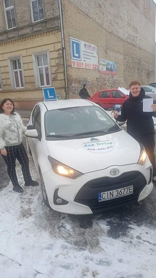 Uśmiechnięta kobieta w futrzanej kurtce i kursant z prawem jazdy pozują obok białej Toyoty Yaris z naklejką 'Eco Drive' i niebieskim 'L' na dachu, na tle budynku z szyldem.