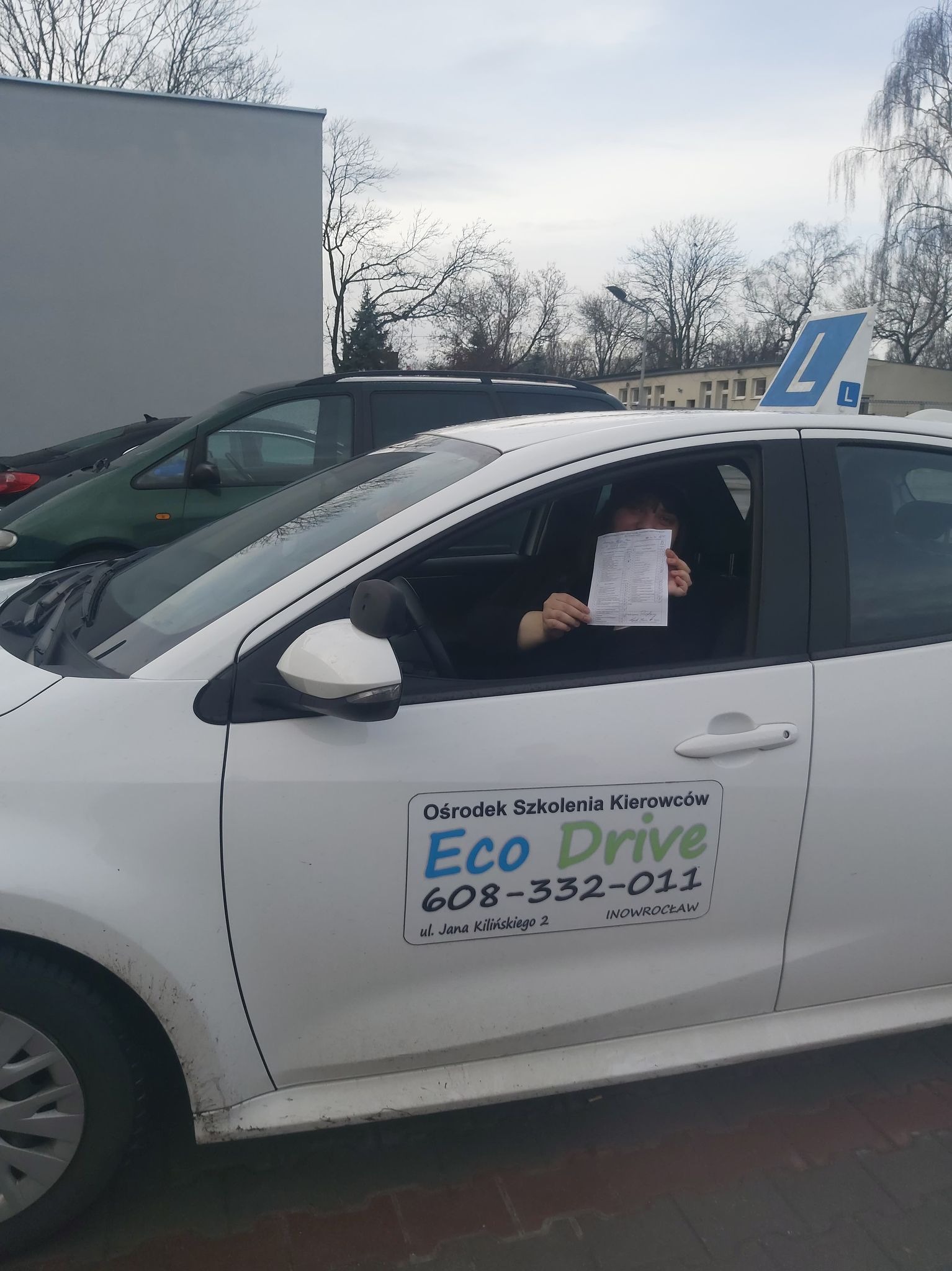 Osoba siedząca w samochodzie z symbolem 'L' na dachu, trzymająca w rękach dokument, na karoserii naklejka z nazwą 'Eco Drive' i numerem telefonu.