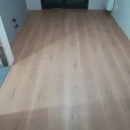 Jasne panele podłogowe ułożone w pokoju z szarą ścianą i czarnymi drzwiami balkonowymi, widoczne gniazdka elektryczne na ścianie.