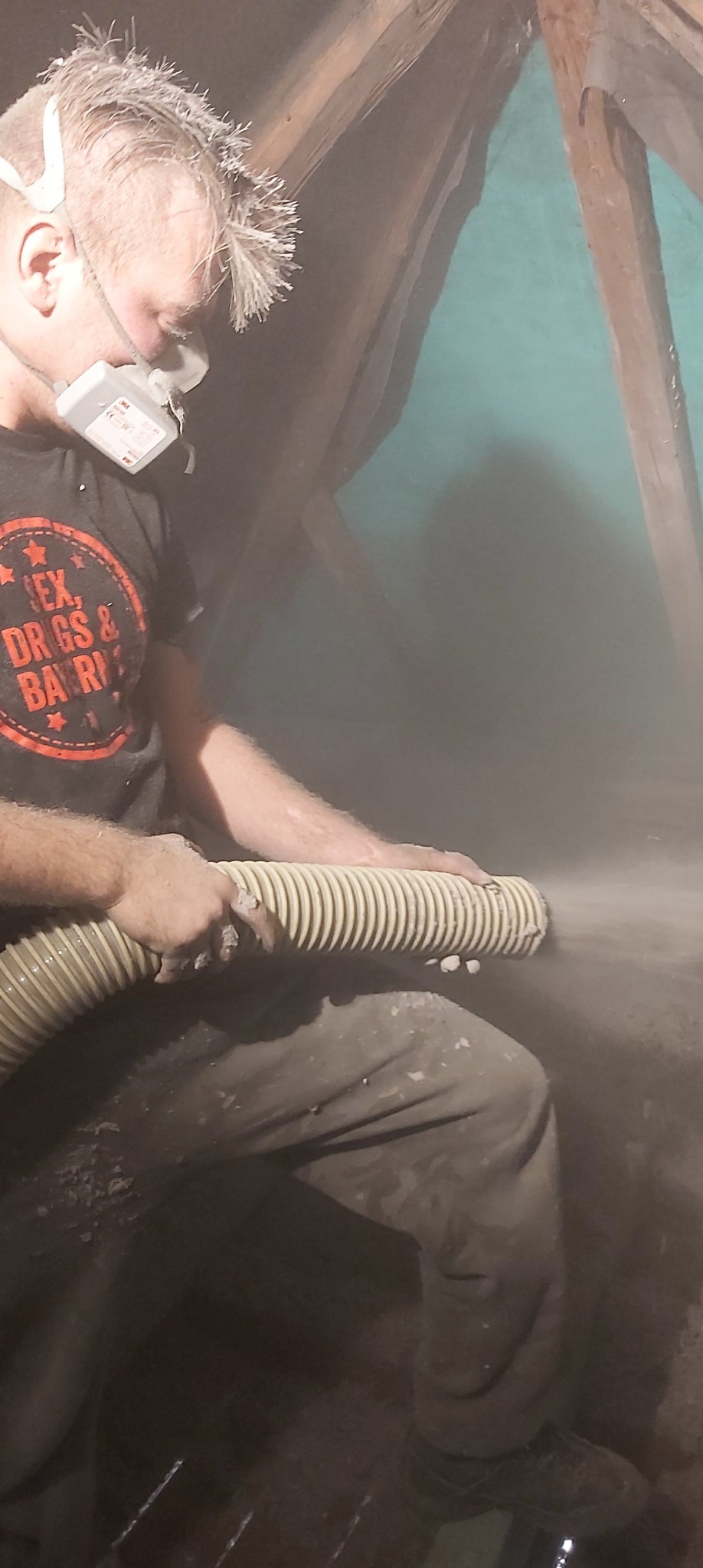 Pracownik w masce ochronnej aplikuje izolację za pomocą węża w przestrzeni poddasza z widocznymi elementami drewnianej konstrukcji dachu.