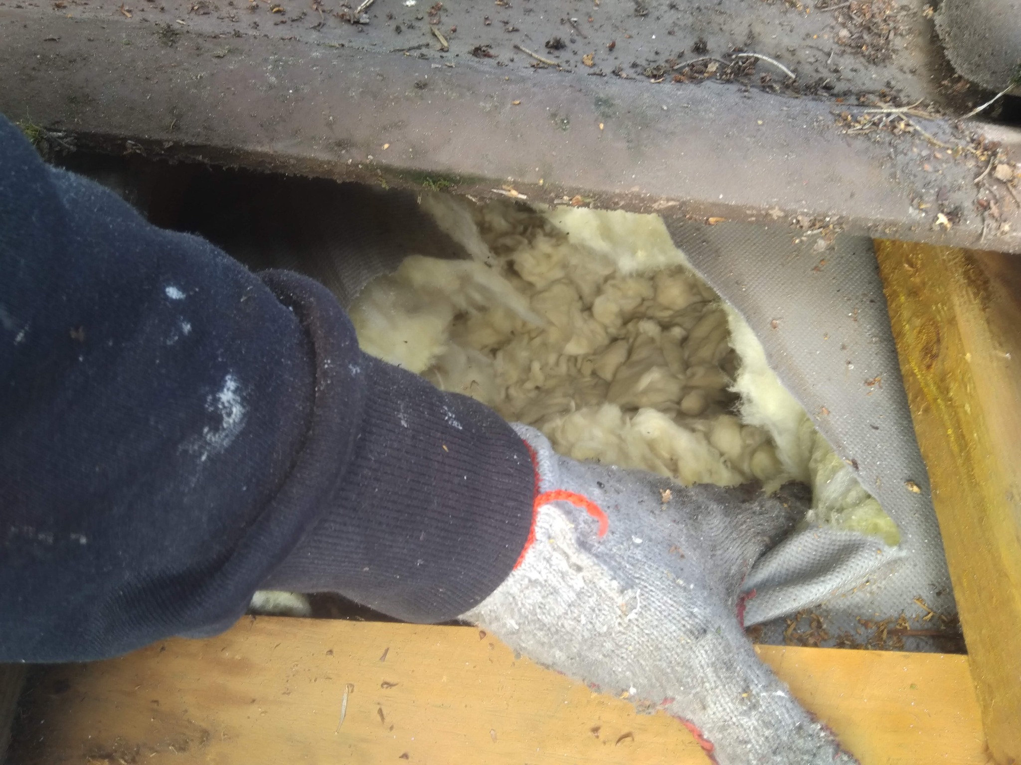 Instalacja izolacji z wełny mineralnej na poddaszu, widoczne drewniane elementy konstrukcji i ręka w roboczej rękawicy z pomarańczową nitką.