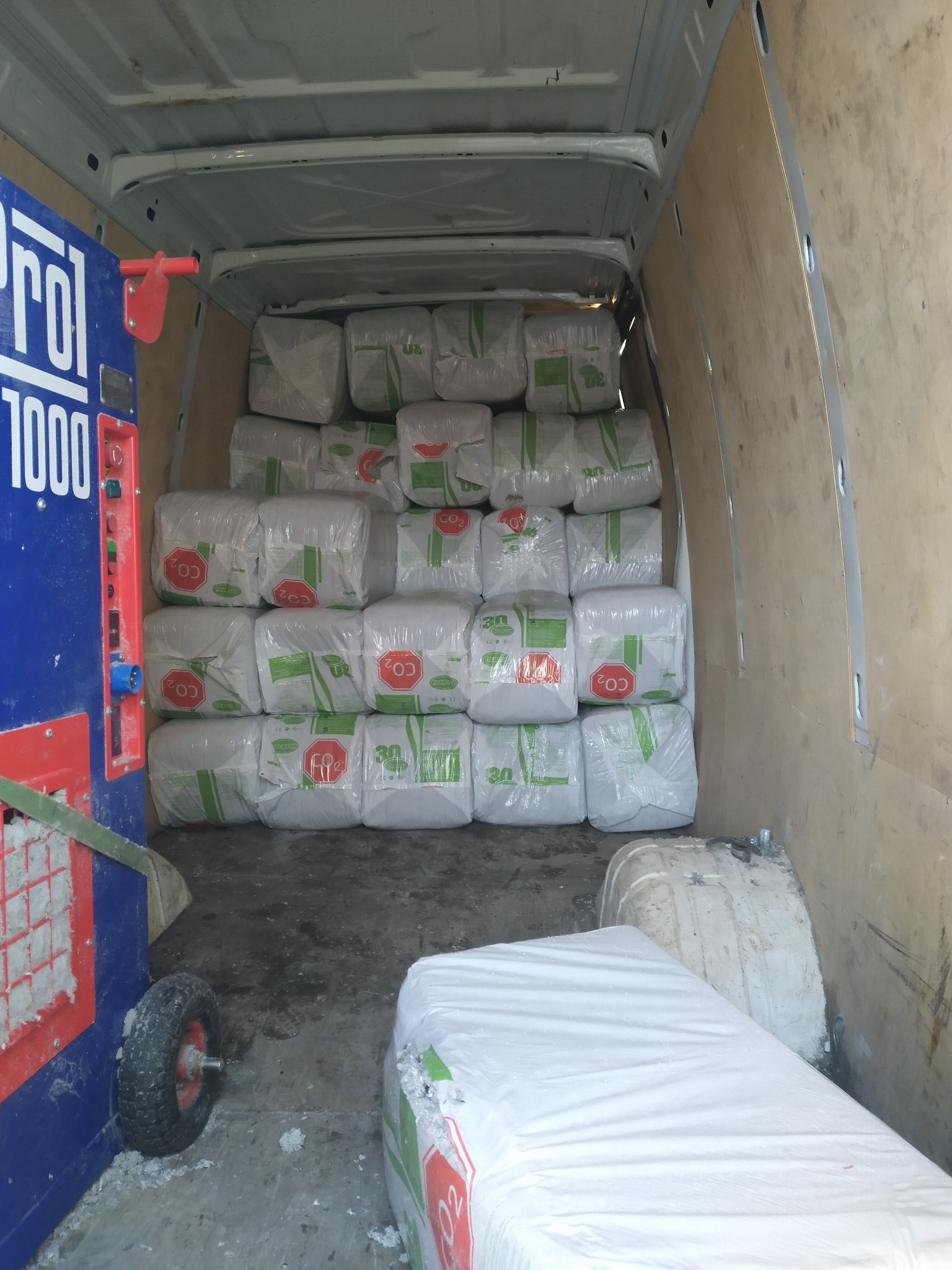 Wnętrze furgonetki wypełnionej paczkami izolacji z oznaczeniami CO2, obok maszyna Pro 1000 do aplikacji, widok z tyłu pojazdu.