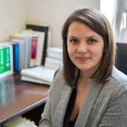 Portret młodej kobiety w marynarce w kratę siedzącej przy biurku z książkami prawniczymi w tle, patrzącej w obiektyw.