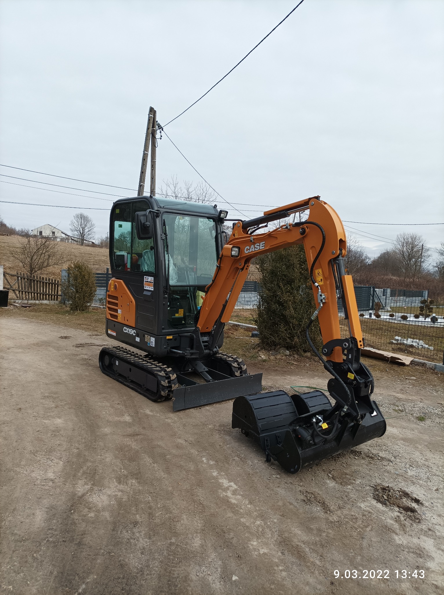 Kompaktowa, pomarańczowo-czarna koparka CASE CX19C na gąsienicach, stojąca na ubitej ziemi z dwoma czarnymi łyżkami z przodu, w tle ogrodzenie i słup elektryczny.