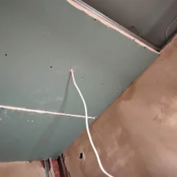 Montaż płyty gipsowo-kartonowej na suficie z wystającym przewodem elektrycznym, widoczne metalowe profile i surowa ściana w tle.