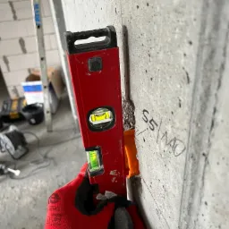 Czerwona poziomica przykładana do szarej, surowej ściany z widocznym otworem na puszkę elektryczną i odręcznym napisem 'S5/MO'