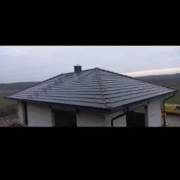 Nowoczesny dom jednorodzinny w trakcie budowy z ciemnoszarą dachówką ceramiczną i zamontowanymi rynnami, widoczne elementy konstrukcyjne i krajobraz w tle.