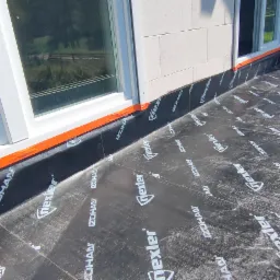 Izolacja przeciwwodna z papy na fundamencie balkonu przy ścianie z bloczków i oknach PCV, zabezpieczona pomarańczową taśmą.