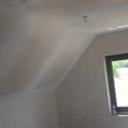 Poddasze w trakcie wykańczania, ściany i skośny sufit pokryte warstwą gładzi szpachlowej, widoczne kable oświetleniowe wystające z sufitu, okno z butelką wody na parapecie.