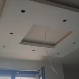 Wykończony sufit podwieszany z wnęką i otworami na oświetlenie punktowe, widoczne kable, w tle okno z widokiem na zabudowania.