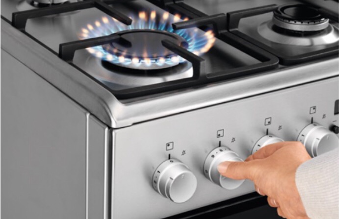 Włączony palnik gazowy w nowoczesnej, srebrnej kuchence; dłoń reguluje pokrętło.