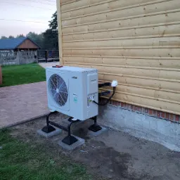 Zewnętrzna jednostka pompy ciepła York zamontowana na metalowej konstrukcji, z widocznym podłączeniem elektrycznym i rurami chłodniczymi, na tle drewnianej elewacji domu i trawnika.