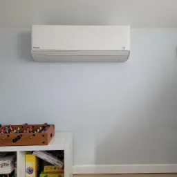 Biały klimatyzator Panasonic zamontowany na jasnoniebieskiej ścianie, poniżej biała półka z grami planszowymi i piłkarzykami, jasna podłoga.