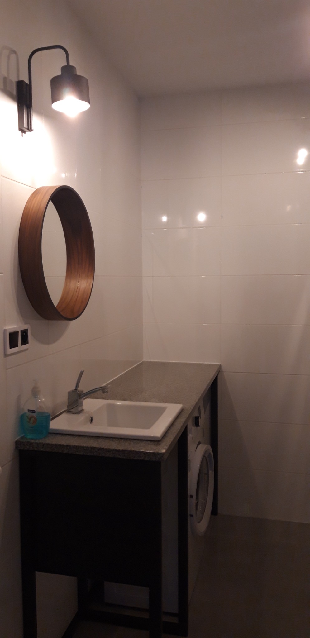 Nowoczesna łazienka z białymi płytkami, okrągłym drewnianym lustrem, umywalką wbudowaną w blat i pralką ukrytą pod blatem, oświetlona lampą ścienną.