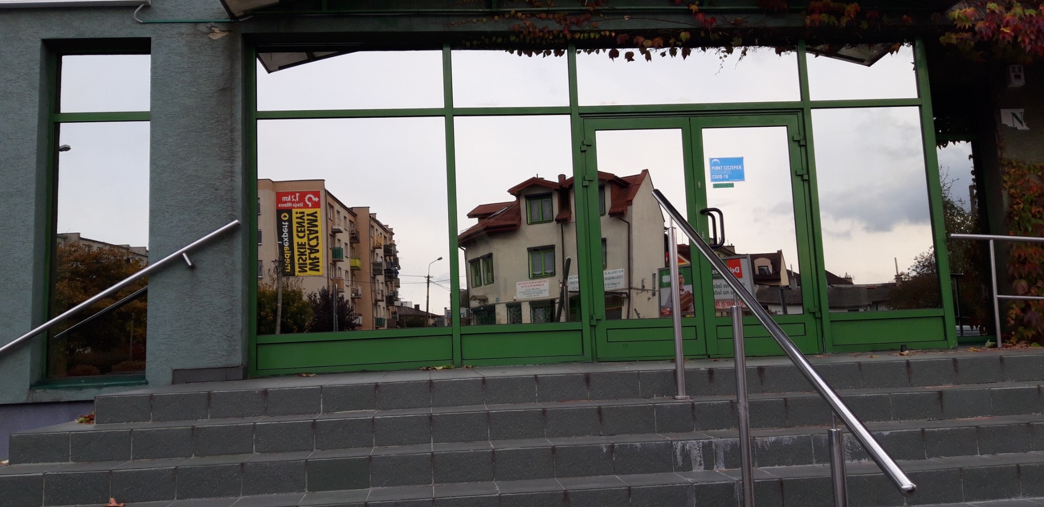 Szklane drzwi wejściowe do budynku z zieloną ramą, na których widoczne jest odbicie pobliskich budynków i nieba, z widocznymi schodami i metalową barierką.