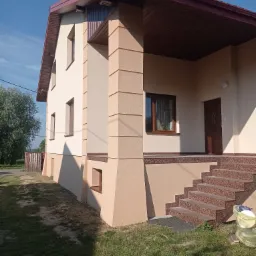 Dwukondygnacyjny dom jednorodzinny z beżową elewacją i brązowym dachem, widoczne schody wejściowe z brązowego granitu, trawnik z workami śmieci obok schodów.