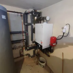 Nowoczesny, biały piec gazowy z czerwonym zbiornikiem wyrównawczym, podłączony do instalacji grzewczej z izolowanymi rurami, widoczny zbiornik na wodę i wiadro z wodą na betonowej podłodze.