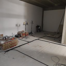 LMElectric Leszek Maciejewski - Wnętrze w trakcie remontu z widocznymi przewodami w listwach podłogowych, materiałami izolacyjnymi ROCKWOOL, narzędziami i lampami budowlanymi.