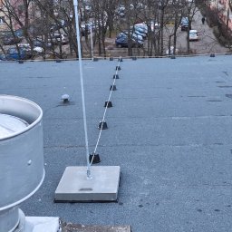 LMElectric Leszek Maciejewski - System ochrony odgromowej na dachu budynku, widoczne betonowe podstawy i przewody odprowadzające, w tle drzewa i budynki mieszkalne.