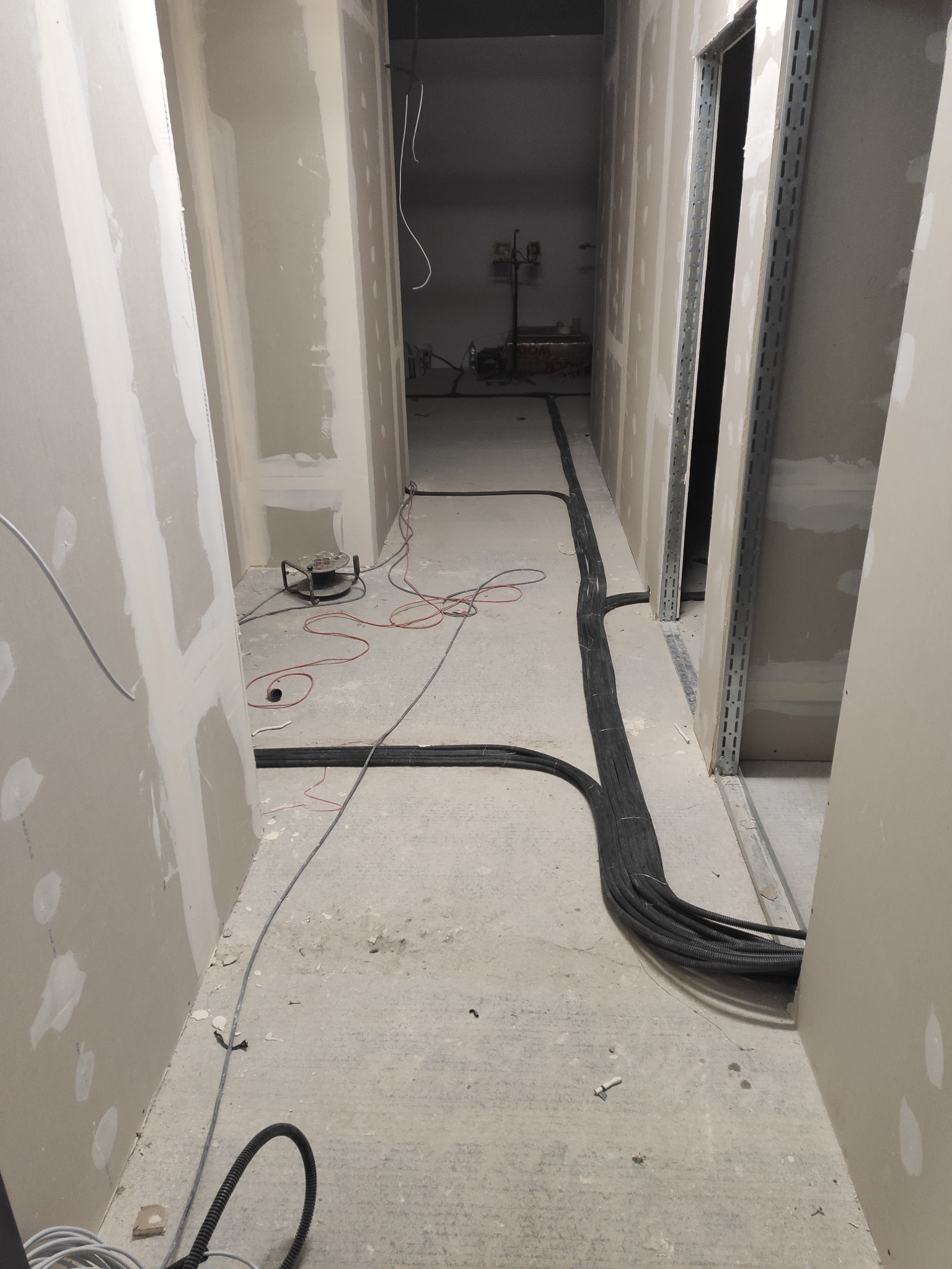 Korytarz w trakcie remontu z widocznymi przewodami elektrycznymi ułożonymi wzdłuż ściany, ściany w stanie surowym, widoczne puszki instalacyjne i rozwijak kabli.