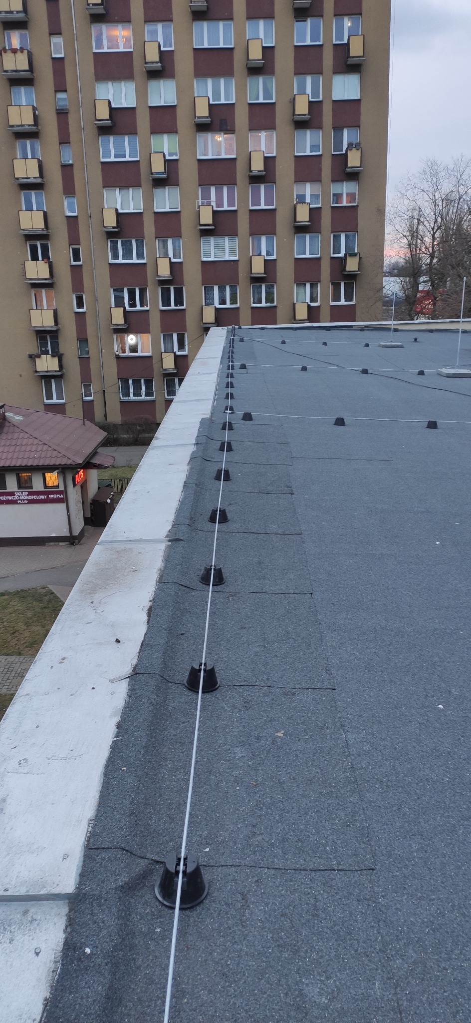 Dach budynku pokryty papą z zamontowaną instalacją odgromową, widoczne czarne uchwyty i biały przewód odprowadzający, w tle blok mieszkalny i sklep.