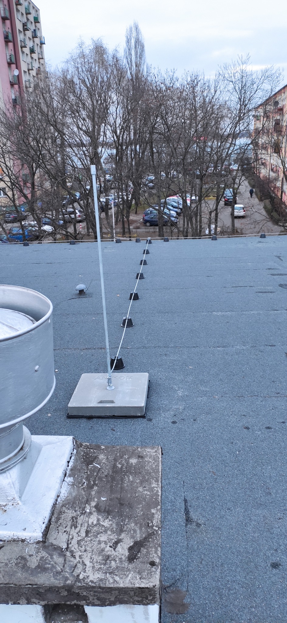 System ochrony odgromowej na dachu budynku, widoczne betonowe podstawy i przewody odprowadzające, w tle drzewa i budynki mieszkalne.