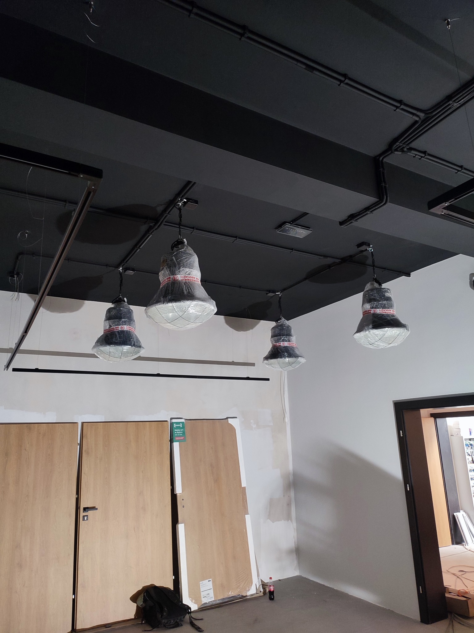 Cztery lampy przemysłowe, zabezpieczone folią ochronną, zwisają z czarnego sufitu z widocznymi instalacjami elektrycznymi. W tle surowe ściany i drzwi, sugerujące etap wykończeniowy.