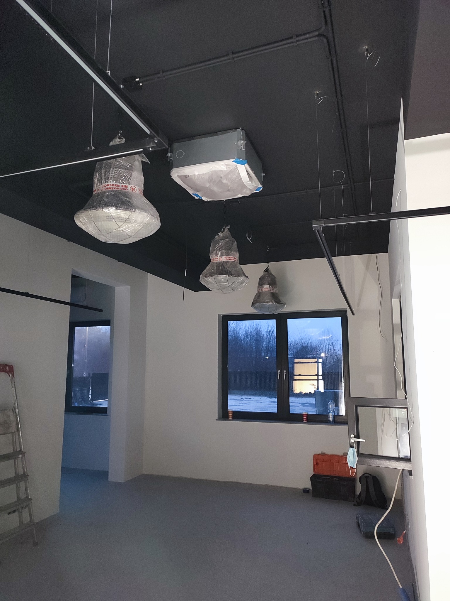 Wnętrze lokalu w trakcie prac wykończeniowych, widoczne podwieszane oprawy oświetleniowe w folii ochronnej, instalacje na suficie, okna z widokiem na zewnątrz, drabina i skrzynki narzędziowe.